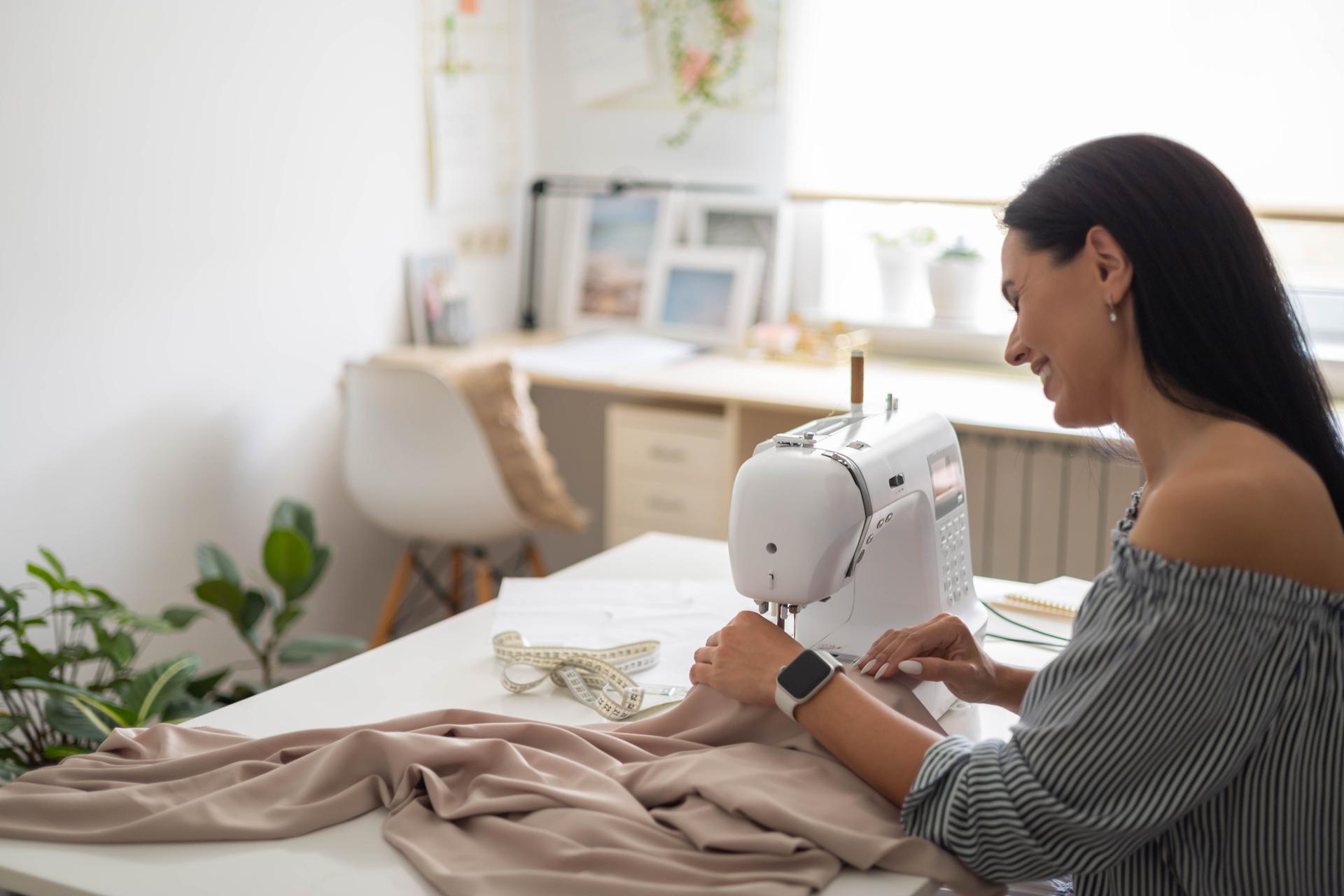 Smiling woman skilled tailor clothes manufacture process on sewing machine posing at workplace